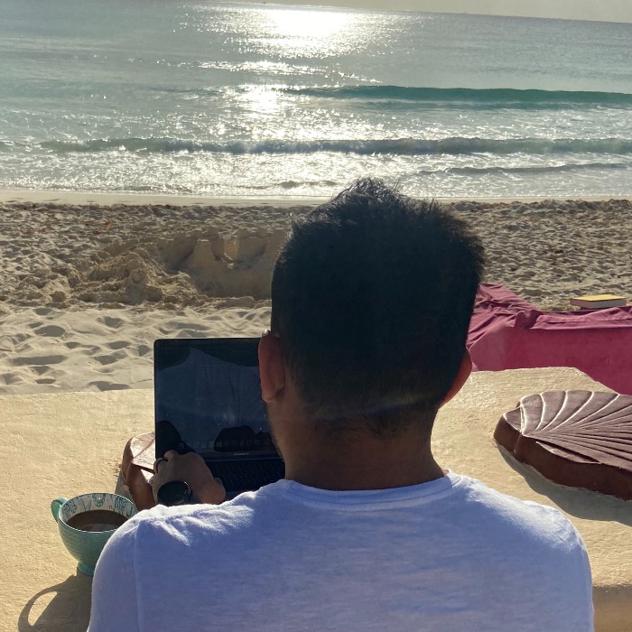 An employee hard at work on the beach: sun shining, waves glistening, laptop open, coffee fresh.