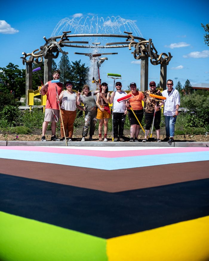 The proud artists and designers standing with their mural (the Progress Pride Flag painted over a crosswalk) with the iconic Riverfront Park statue in the background.